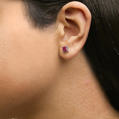 Close-up side view of a model wearing a pair of white metal stud earrings, each featuring a princess-shaped red gemstone set with four prongs.