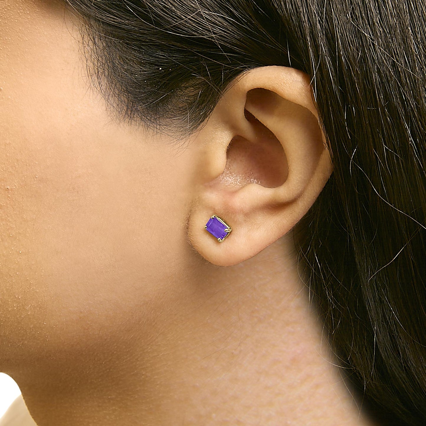 Close-up side view of a model wearing a yellow metal stud earring with a purple emerald-shaped gemstone in a four-prong setting.