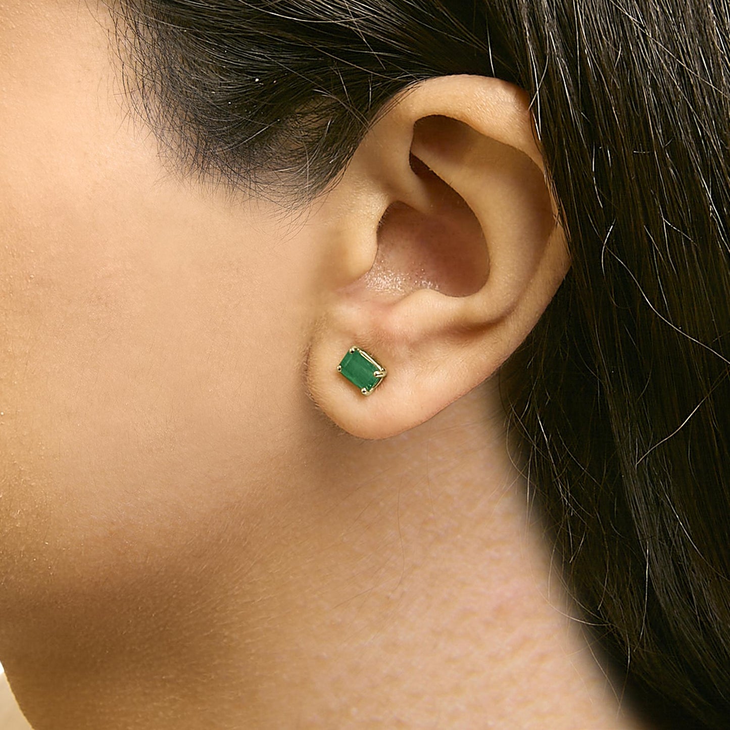 Close-up side view of a model wearing a yellow metal stud earring with a green emerald-shaped gemstone in a four-prong setting.