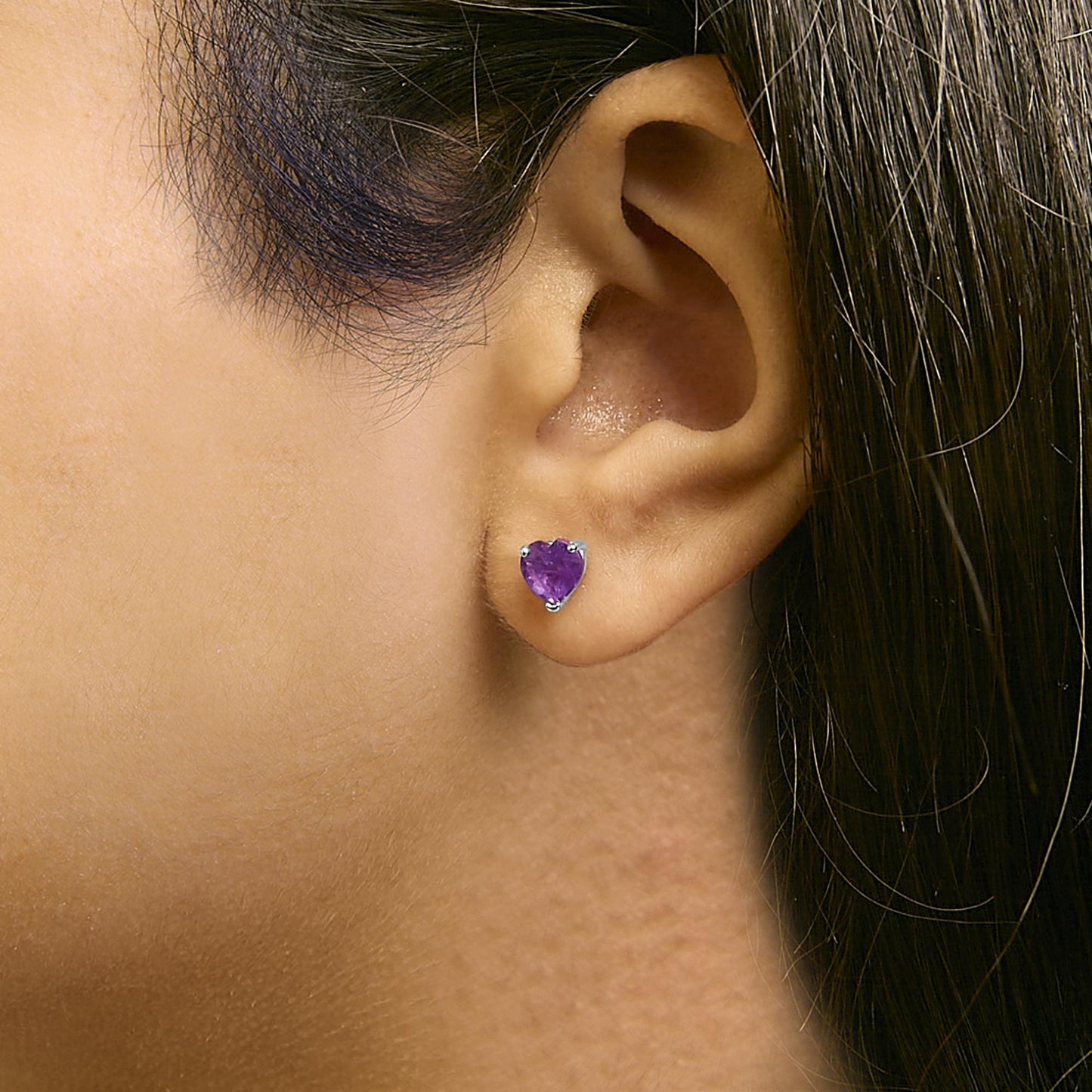 Close-up side view of a model wearing a single white metal earring with a heart-shaped purple gemstone in a 3-prong setting.