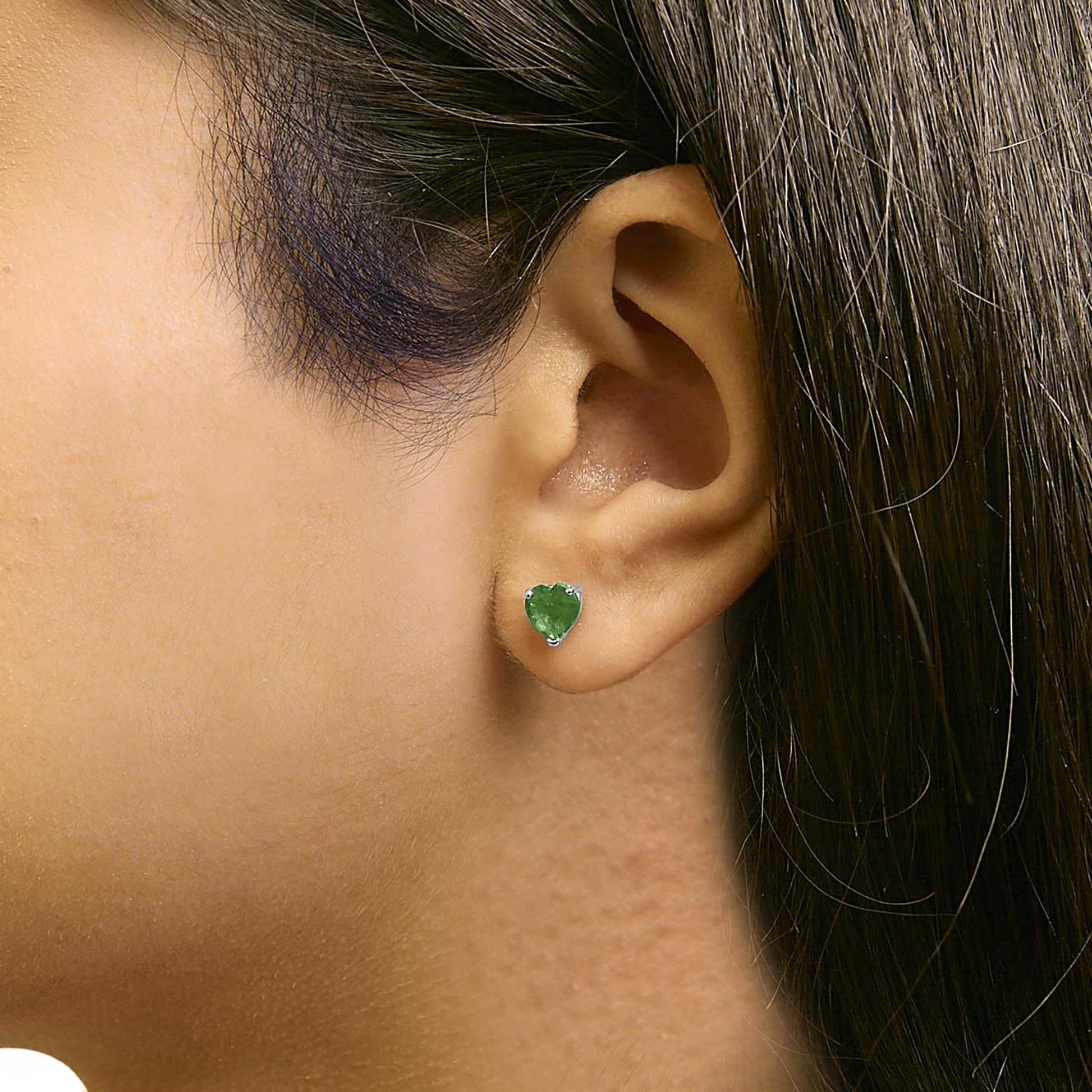 Close-up side view of a white metal earring with a heart-shaped green gemstone in a 3-prong setting, worn on a model's ear.