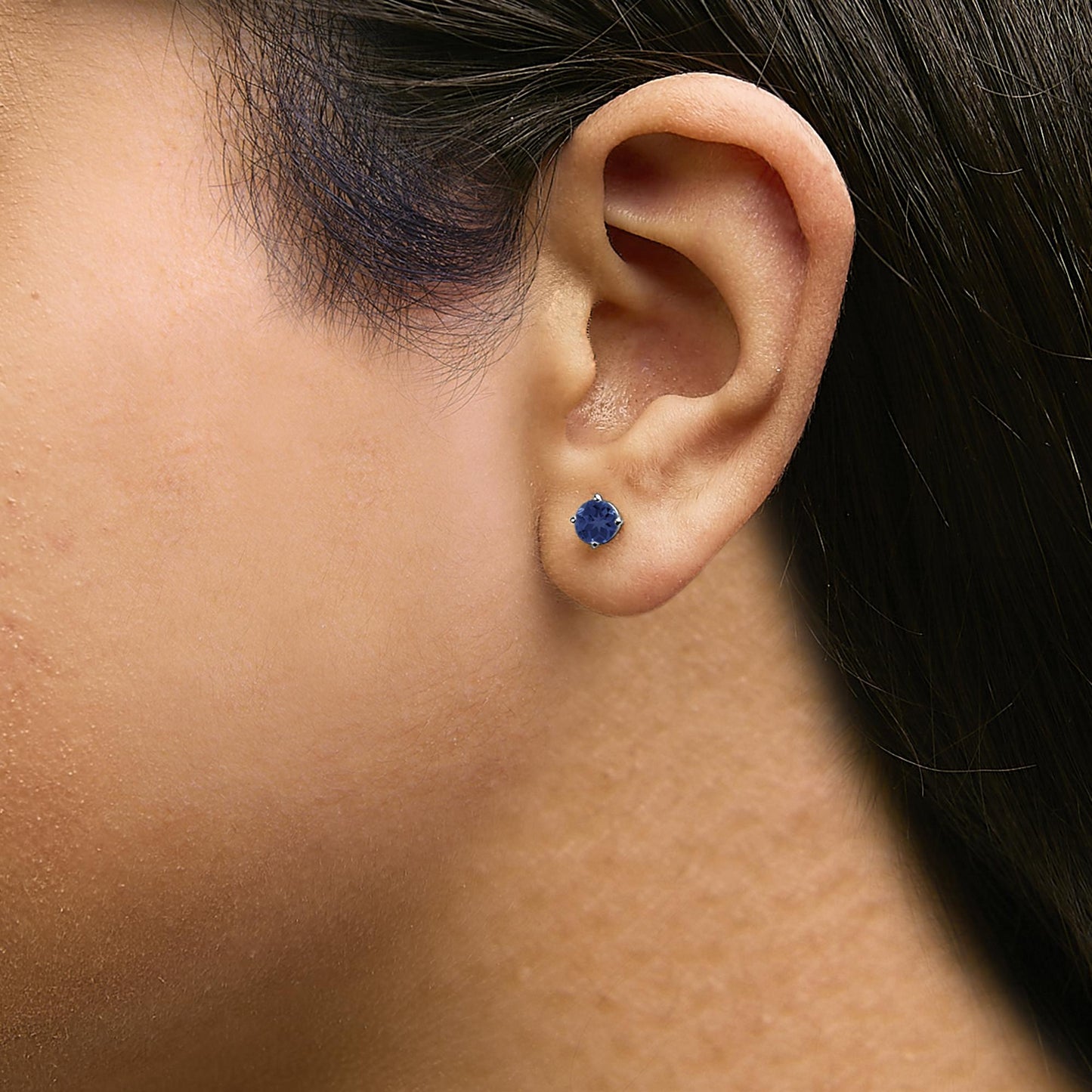 Close-up side view of a model wearing a white metal stud earring with a round blue gemstone in a four-prong setting.