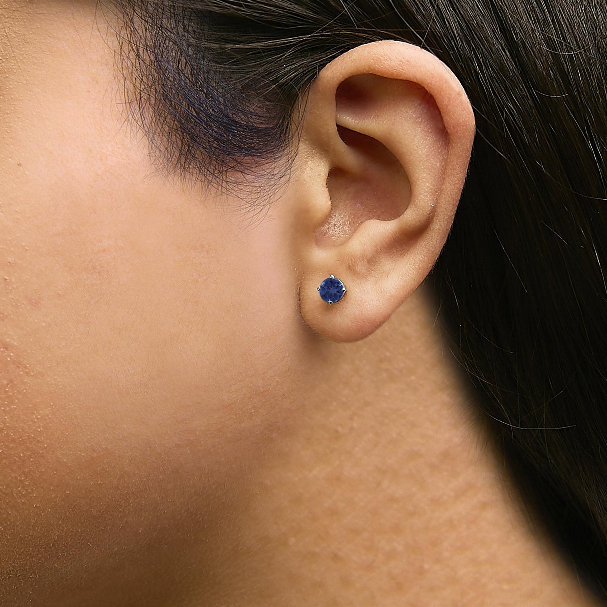 Close-up side view of a model wearing a white metal stud earring with a round blue gemstone in a four-prong setting.