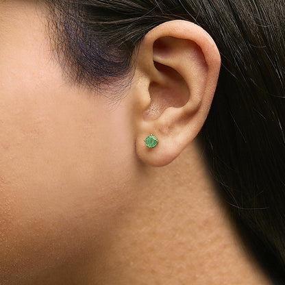 Close-up side view of a model wearing a yellow metal stud earring set with a round green gemstone in a four-prong setting.