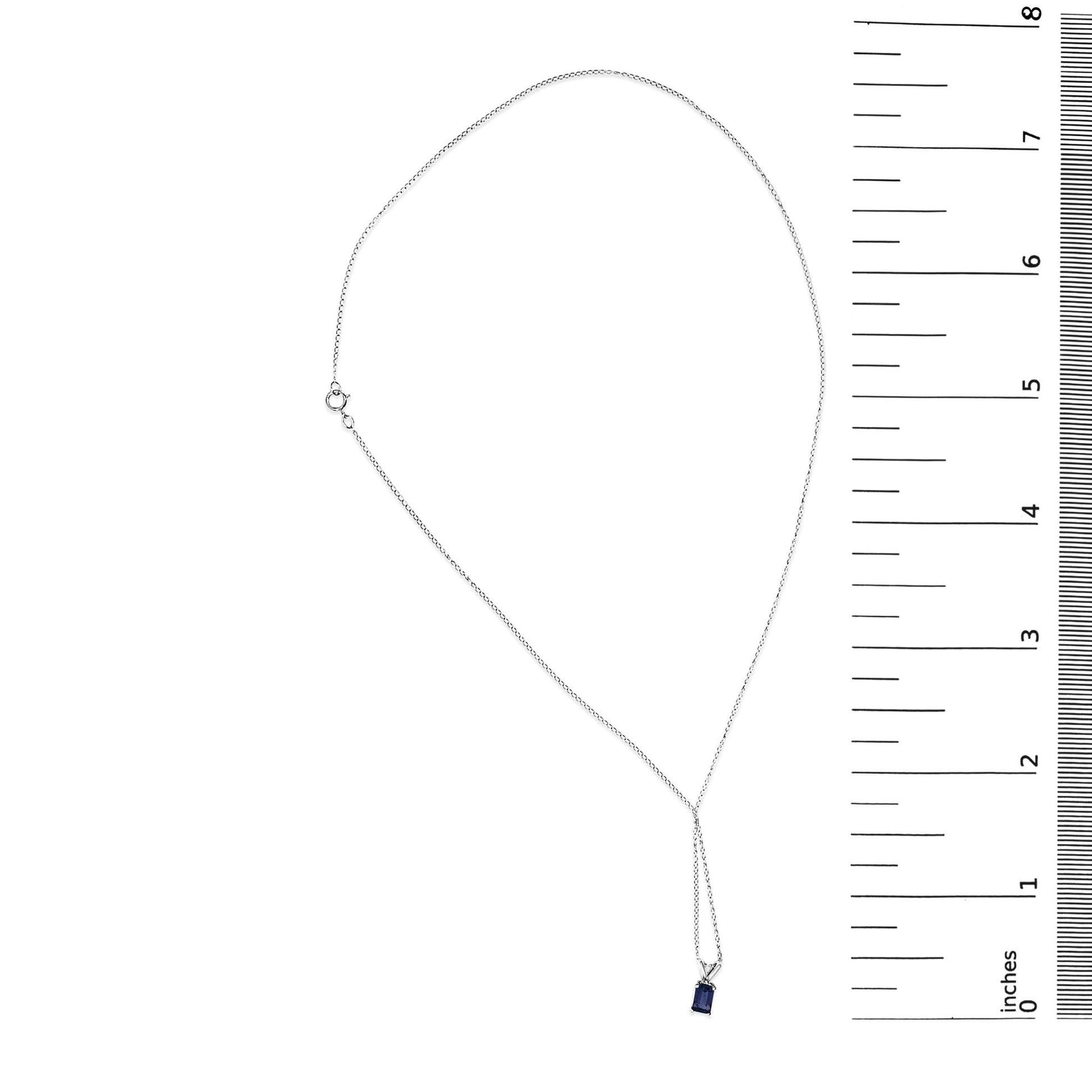 White necklace with a small blue emerald-shaped gemstone pendant set in a 4-prong setting, shown flat on a scale, top-down angle.