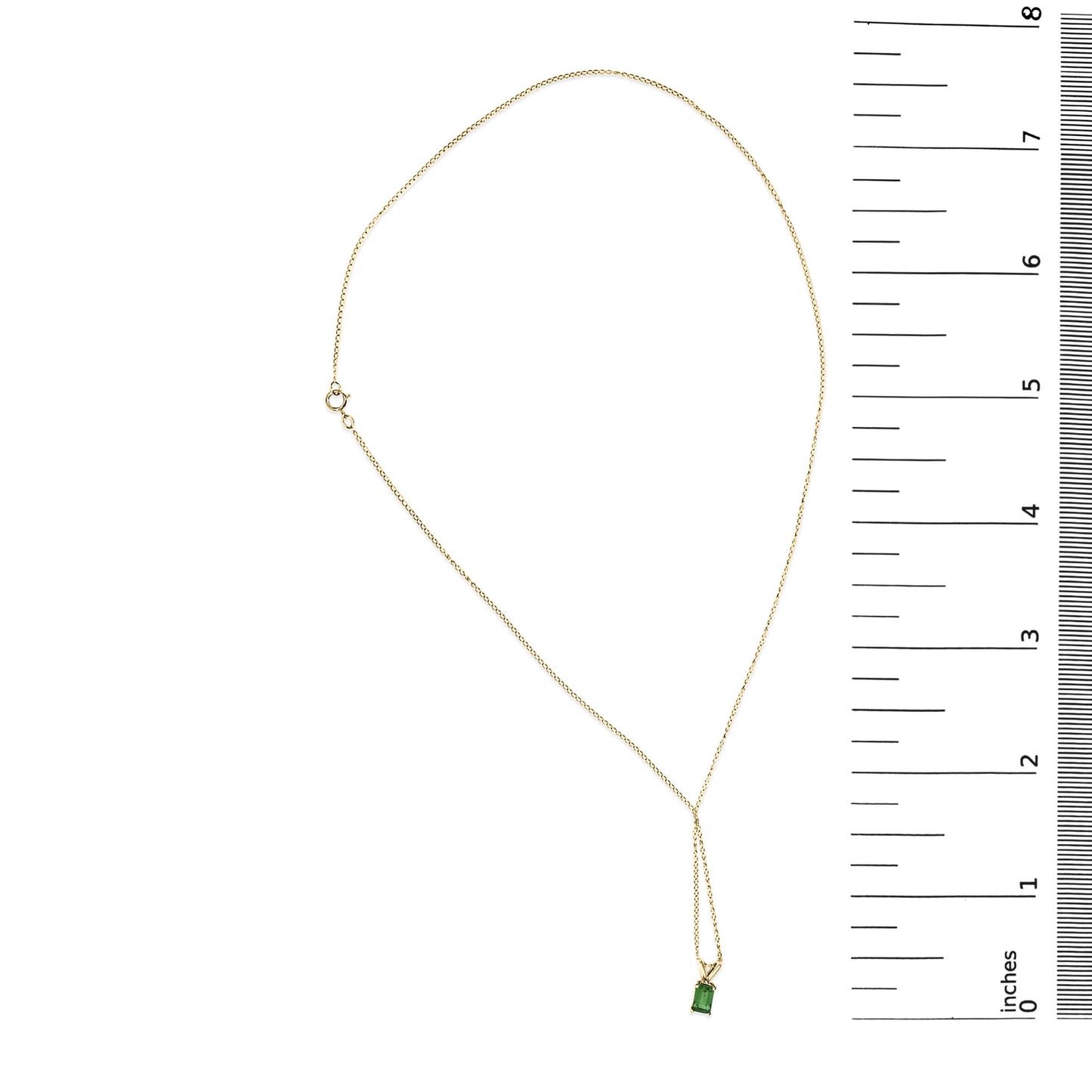 Yellow metal necklace with a chain and a green emerald-shaped gemstone pendant in a four-prong setting, shown on a flat surface next to a ruler scale.