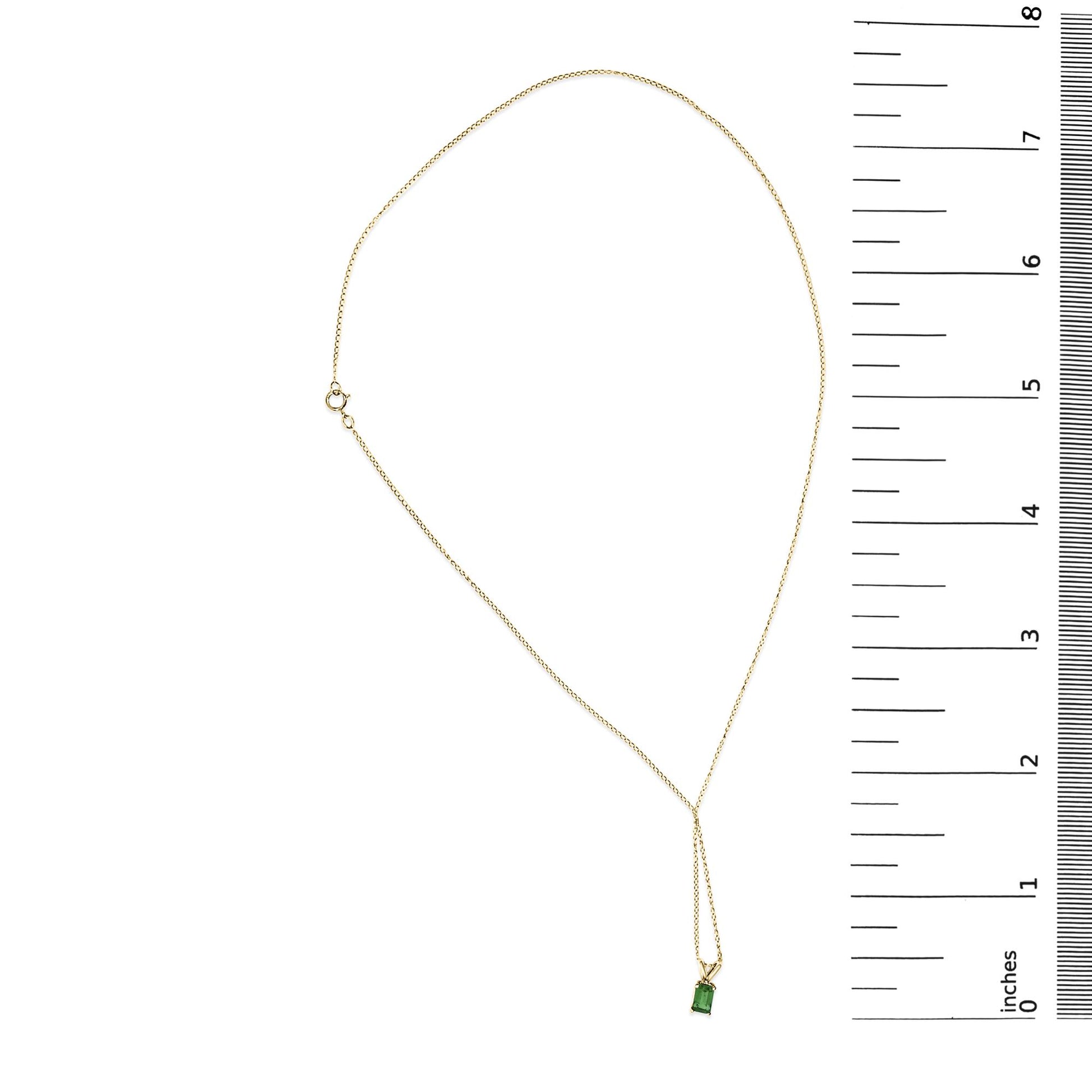 Yellow metal necklace with a chain and a green emerald-shaped gemstone pendant in a four-prong setting, shown on a flat surface next to a ruler scale.