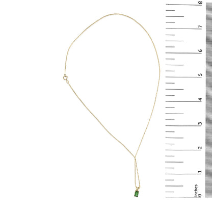 Yellow metal necklace with a chain and a green emerald-shaped gemstone pendant in a four-prong setting, shown on a flat surface next to a ruler scale.