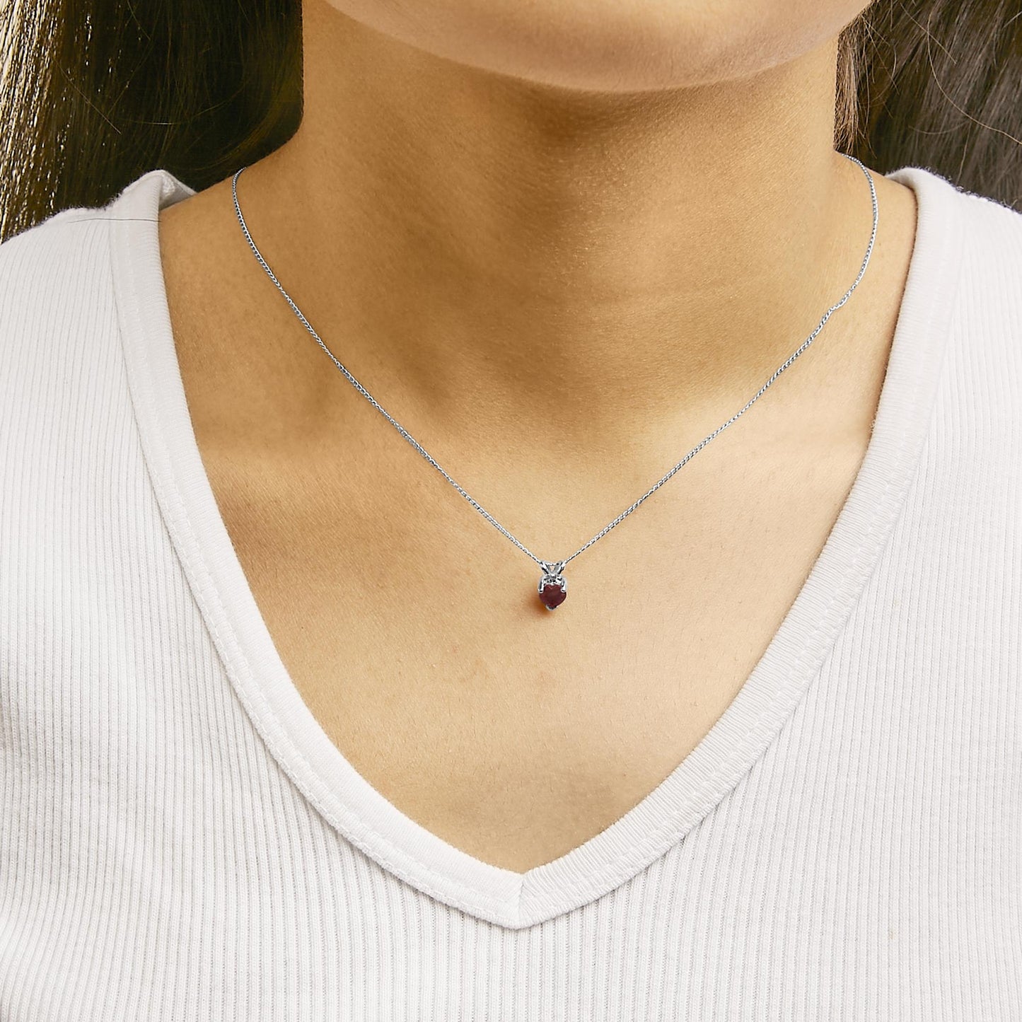 Close-up front view of a model wearing a white metal necklace with a heart-shaped red gemstone pendant held by a three-prong setting.