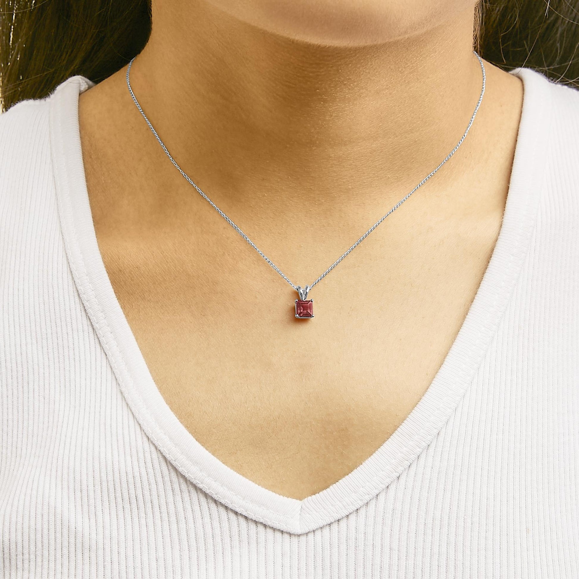 Close-up front view of a white metal necklace with a princess-shaped red gemstone in a four-prong setting, worn on a model.