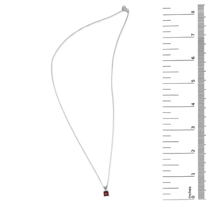White necklace with a princess-shaped red gemstone pendant in a four-prong setting, shown flat on a surface next to a ruler for scale, top-down angle.