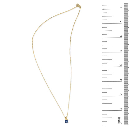 Yellow metal necklace with a princess-shaped blue gemstone pendant in a 4-prong setting, shown flat on a scale from above.