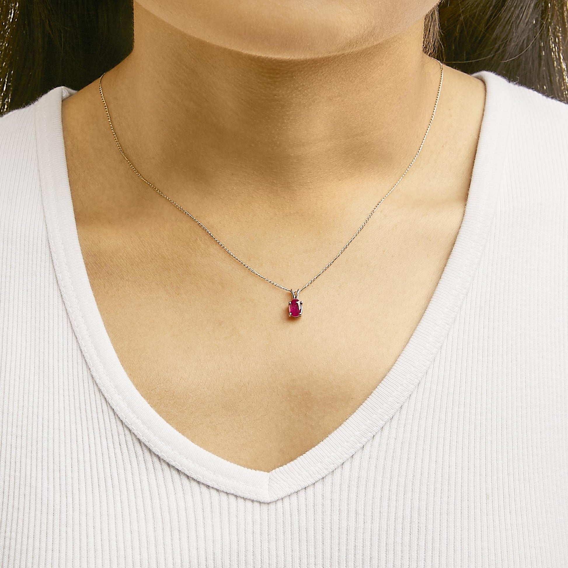 Necklace with a white metal chain featuring a small oval red gemstone pendant in a four-prong setting, shown worn on a model from the front angle.