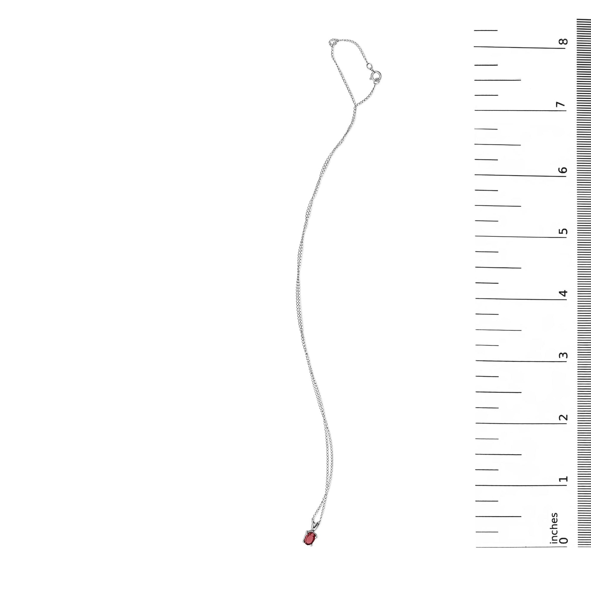 White necklace with an oval red gemstone in a four-prong setting, shown flat next to a ruler from a top-down angle.