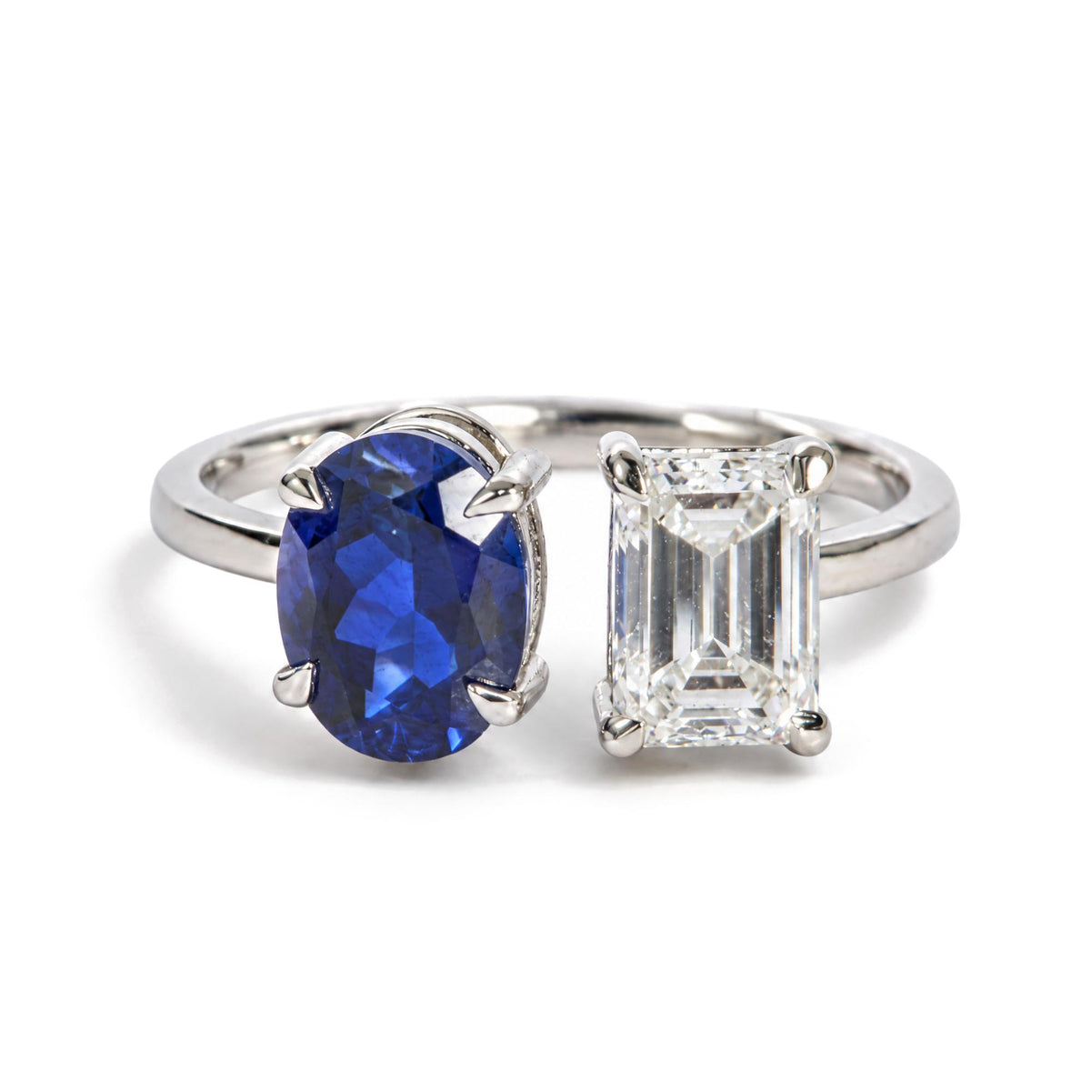 Close-up front angle of a white metal ring shown on a scale, featuring one oval blue gemstone and one emerald-cut white gemstone both in prong settings.