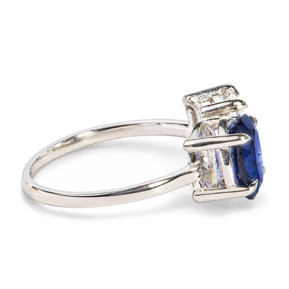 Close-up angled view of a white metal ring featuring a prong-set oval blue gemstone and a prong-set emerald-shaped white gemstone.