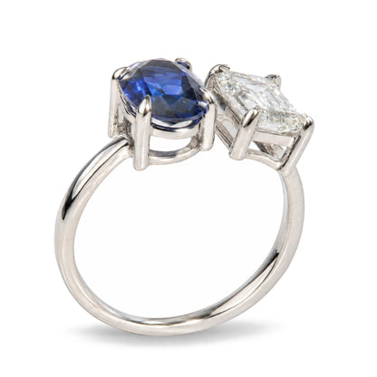 Close-up front angle shot of a white metal ring featuring one oval blue gemstone and one emerald-shaped white gemstone, both in prong settings.