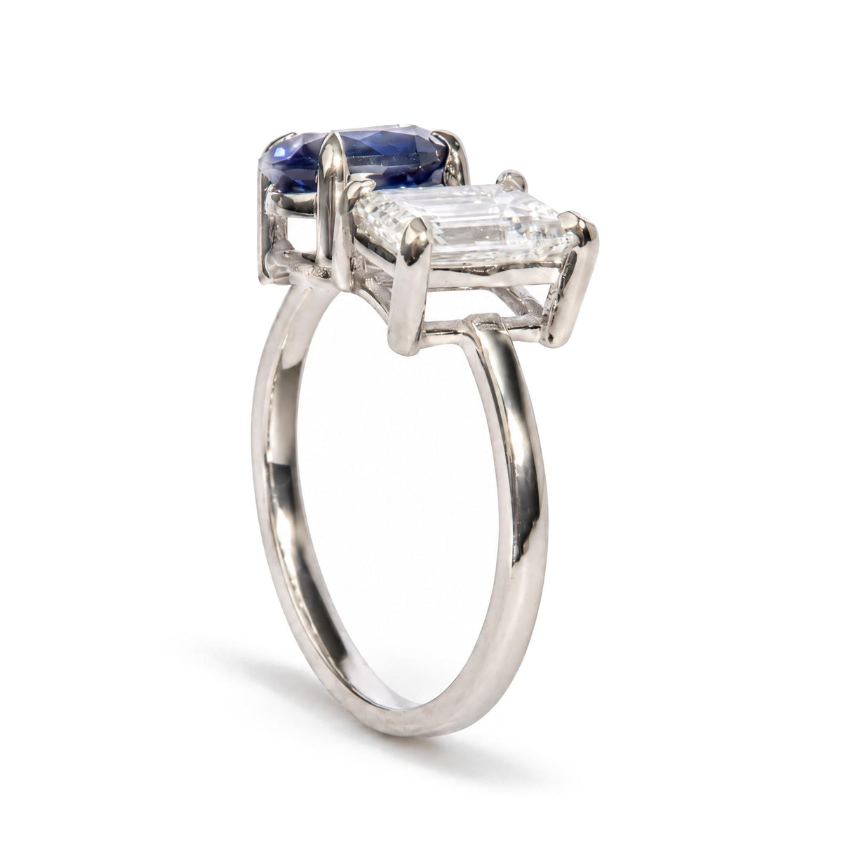 Close-up front angle of a white metal ring featuring a prong-set white emerald-shaped gemstone and a prong-set blue oval-shaped gemstone.