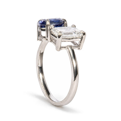 Close-up front angle of a white metal ring featuring a prong-set white emerald-shaped gemstone and a prong-set blue oval-shaped gemstone.
