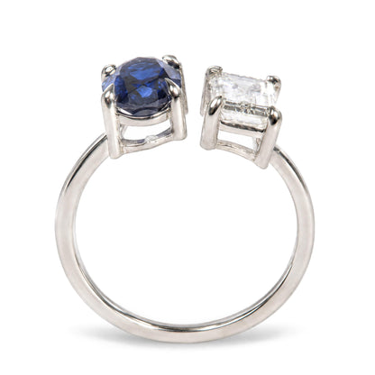 Close-up front angle of a white ring with two prong-set gemstones, one blue oval-shaped and one white emerald-shaped.