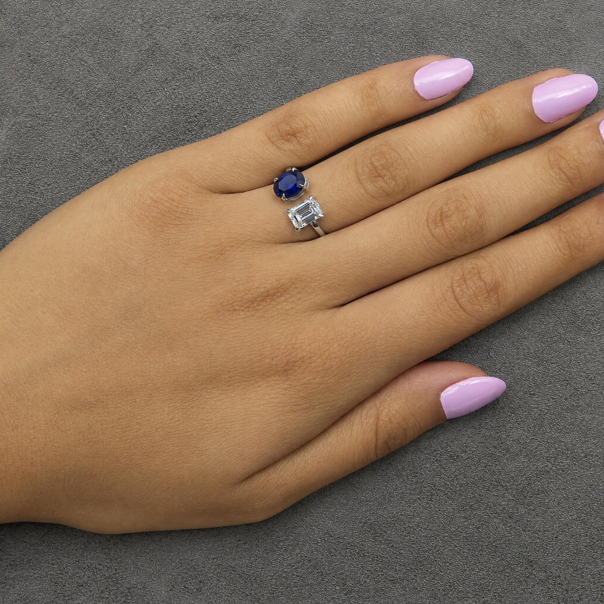 White metal ring with an emerald-shaped white gemstone and an adjacent oval-shaped blue gemstone in prong settings, shown worn on a model's hand from a top angle.