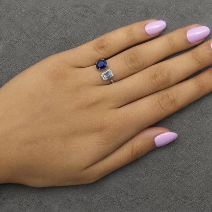 White metal ring with an emerald-shaped white gemstone and an adjacent oval-shaped blue gemstone in prong settings, shown worn on a model's hand from a top angle.