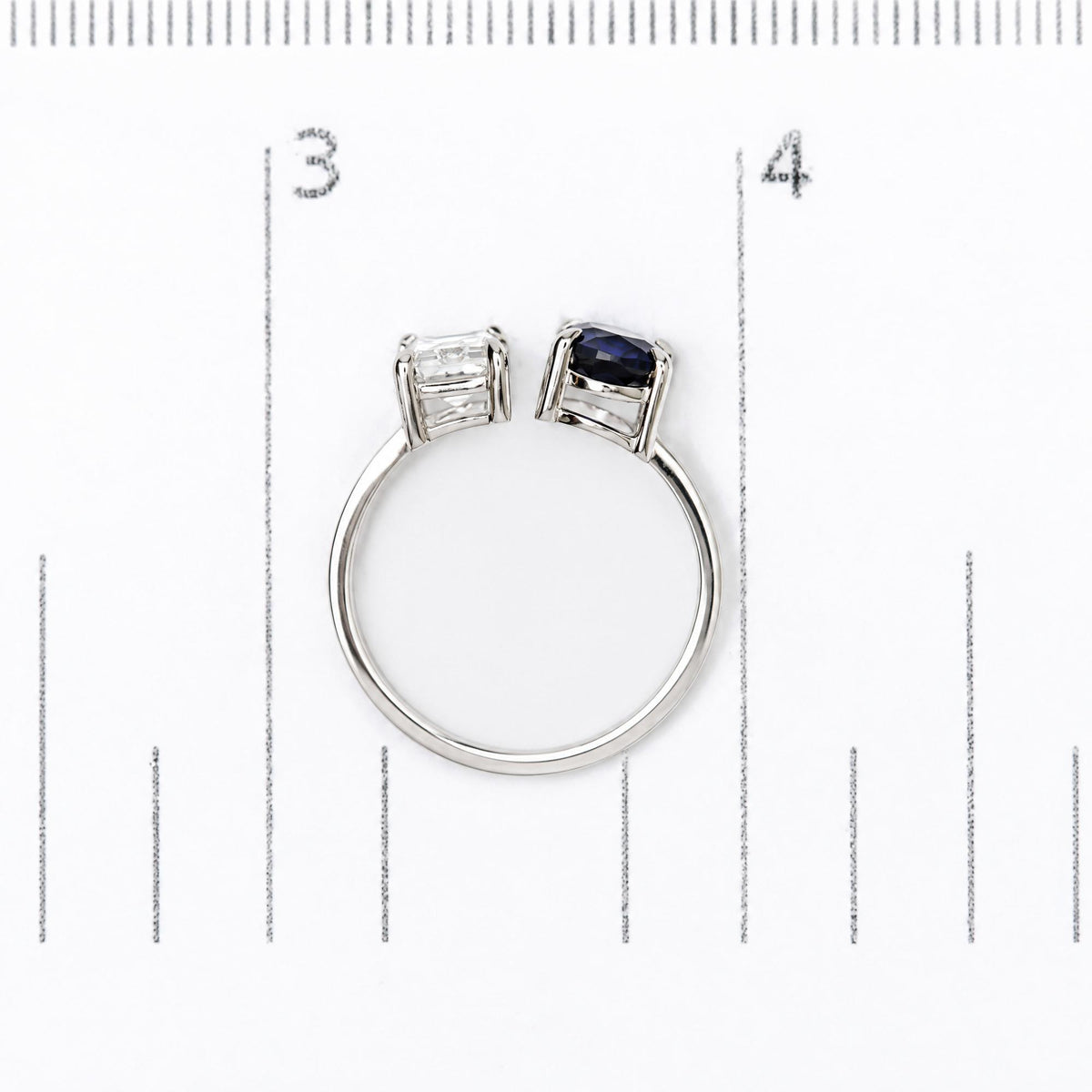 White metal ring shown on a measuring scale, featuring one white emerald-shaped gemstone and one blue oval-shaped gemstone in prong settings, top view.