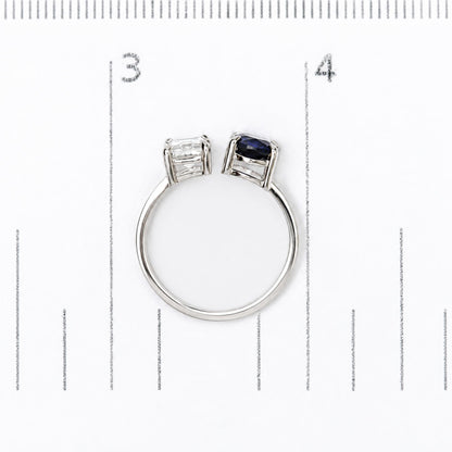 White metal ring shown on a measuring scale, featuring one white emerald-shaped gemstone and one blue oval-shaped gemstone in prong settings, top view.