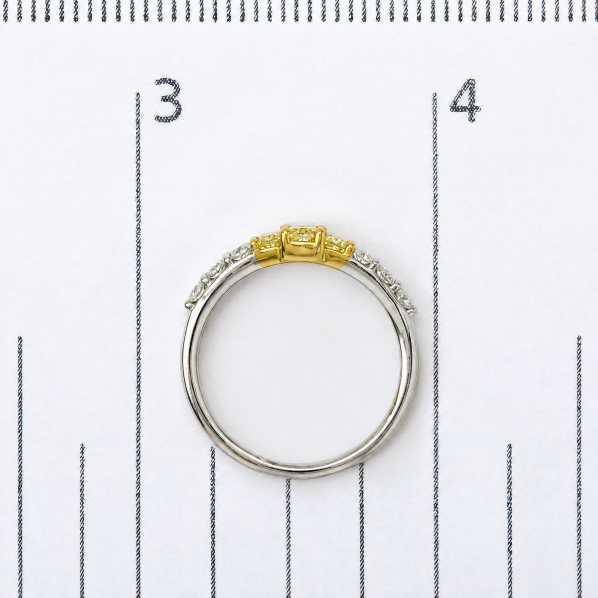 Close-up image of a two-tone ring on a measurement scale, featuring yellow radiant-shaped gemstones in prong settings at the center and white round-shaped gemstones in prong settings along the band, shot from a top-down angle.