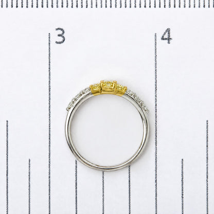 Close-up image of a two-tone ring on a measurement scale, featuring yellow radiant-shaped gemstones in prong settings at the center and white round-shaped gemstones in prong settings along the band, shot from a top-down angle.