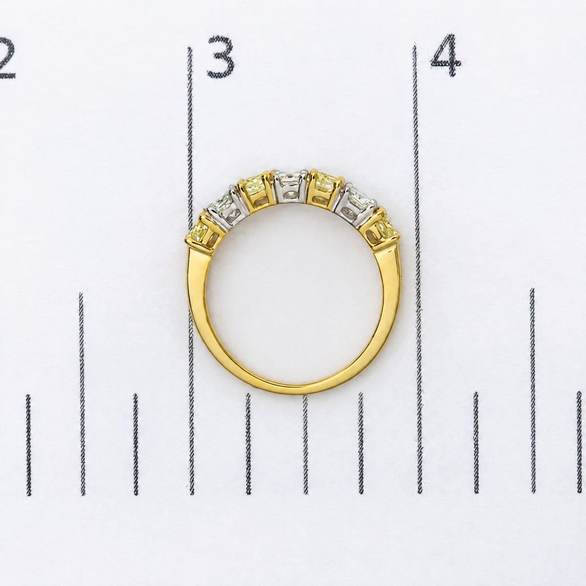 Top-down close-up image of a yellow and white metal ring with alternating yellow and white cushion-shaped gemstones in prong settings, shown on a measurement scale.
