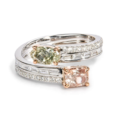 Close-up front angle of two intertwined rings with white and rose metal colors, featuring a cushion-shaped pink gemstone and an oval-shaped green gemstone in prong settings, surrounded by rows of channel-set baguette-shaped white gemstones and prong-set round white gemstones.