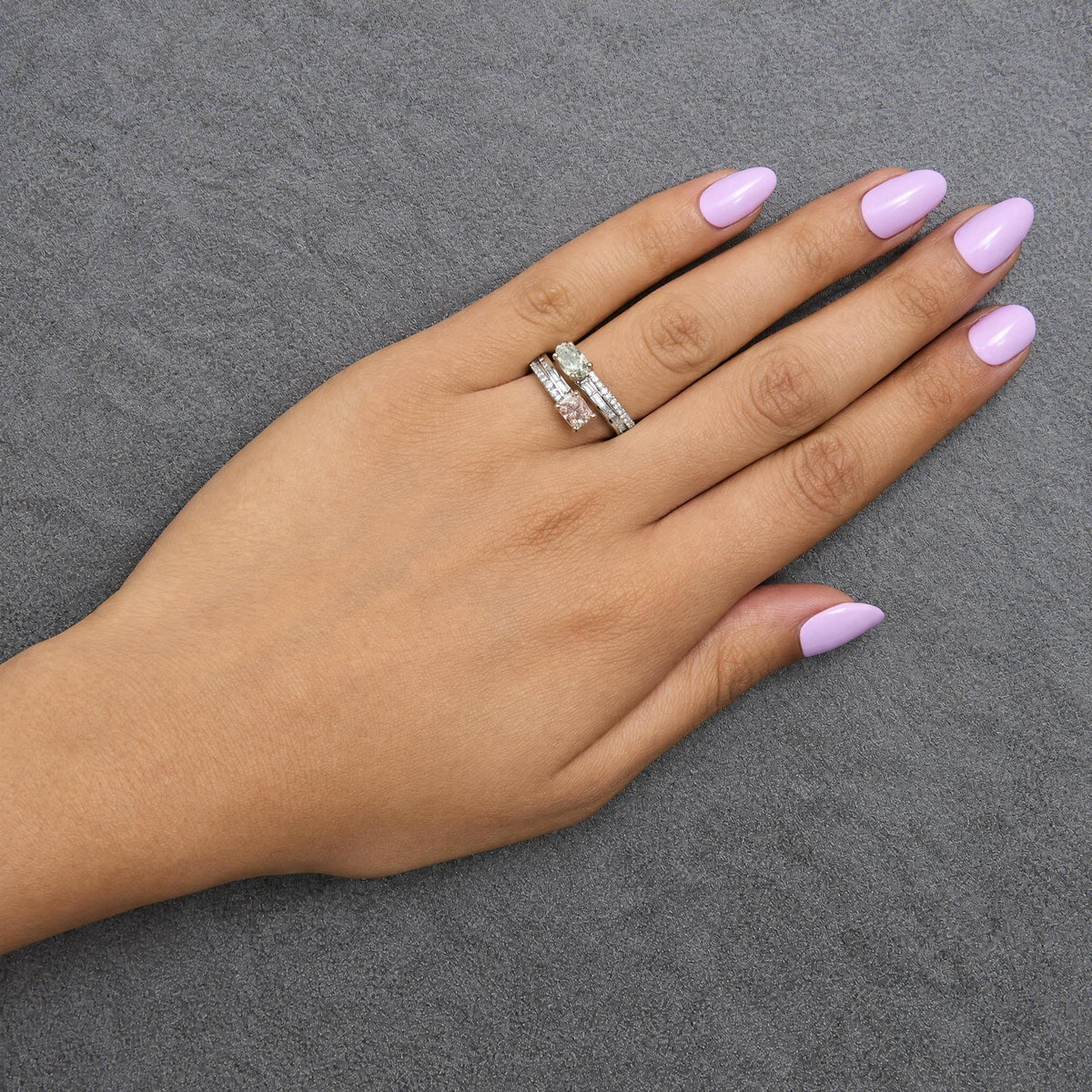Close-up side angle of a model's hand wearing two rings: one featuring a green oval gemstone with a row of white baguette gemstones in a channel setting, and the other with a pink cushion gemstone and round white gemstones in prong settings on a white and rose metal band.