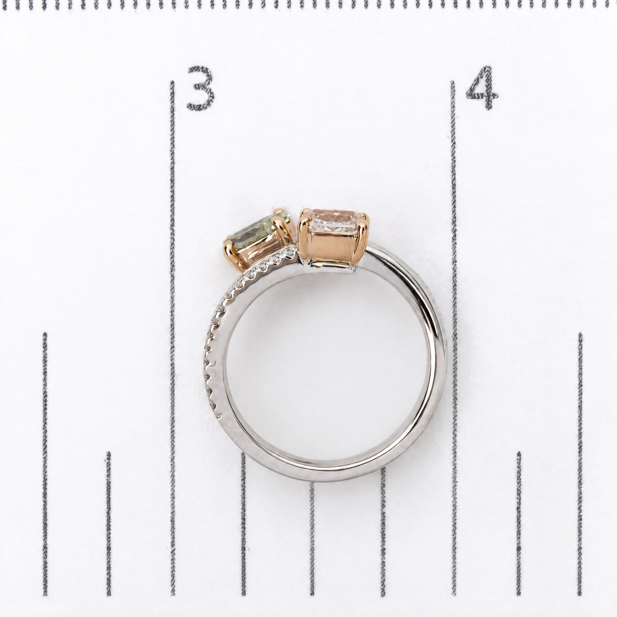 Close-up image of a white and rose metal ring shown on a scale, featuring one pink cushion-shaped gemstone and one green oval-shaped gemstone in prong settings, with multiple white round gemstones in prong settings and white baguette gemstones in channel settings along the band. The ring is viewed from the side angle.