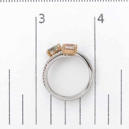 Close-up image of a white and rose metal ring shown on a scale, featuring one pink cushion-shaped gemstone and one green oval-shaped gemstone in prong settings, with multiple white round gemstones in prong settings and white baguette gemstones in channel settings along the band. The ring is viewed from the side angle.