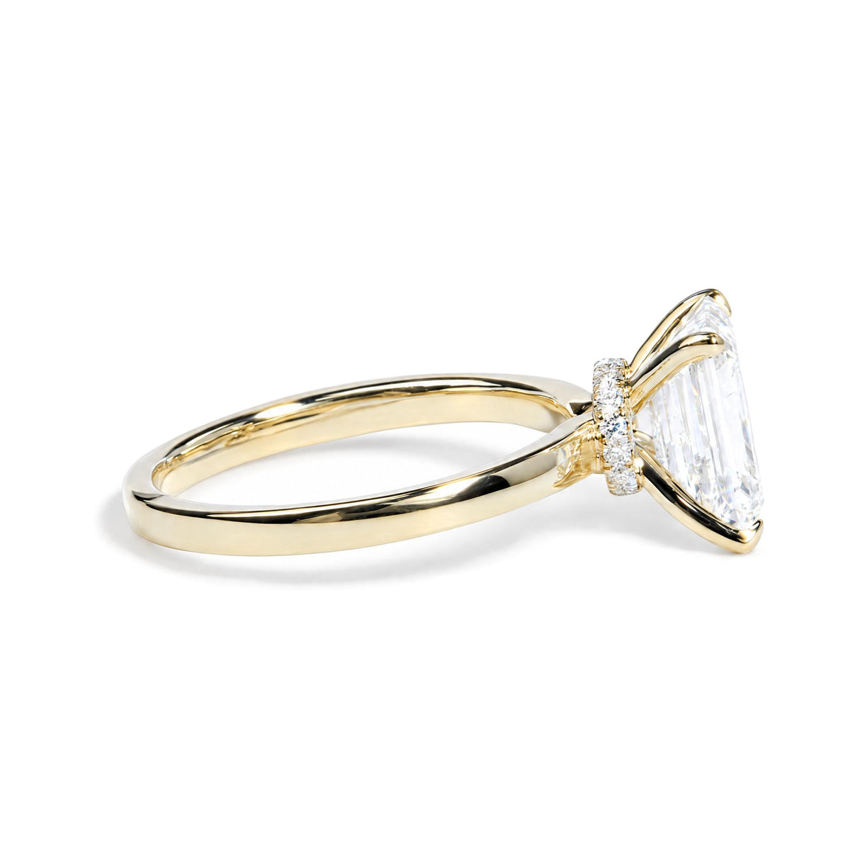 Close-up front angle of a yellow ring set with a large emerald-shaped white gemstone in prong setting, accented by a band of round white gemstones in prong setting.