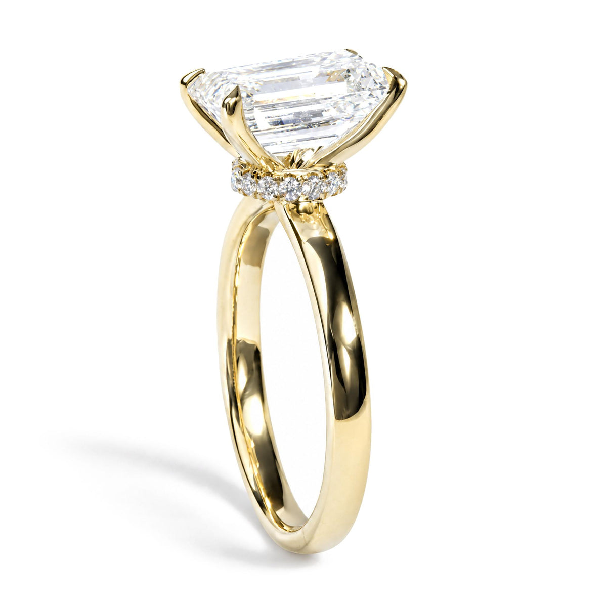 Close-up angled view of a yellow metal ring with a large emerald-shaped white gemstone in a prong setting, surrounded by a band of round white gemstones in prong settings.