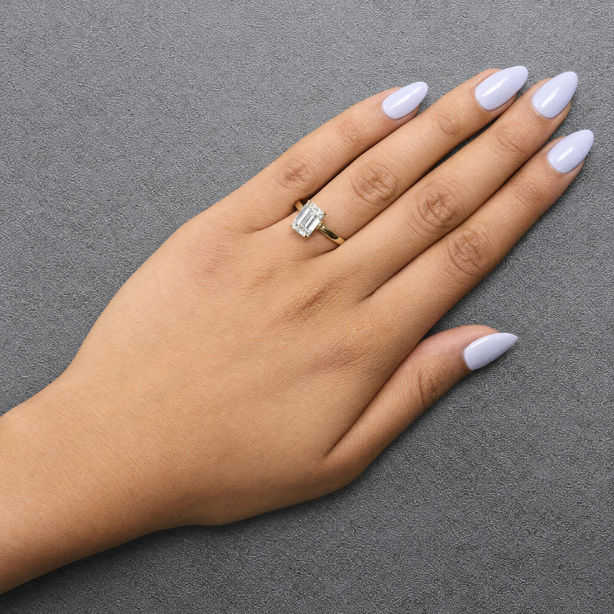 Close-up angled view of a yellow ring with a single emerald-shaped white gemstone and multiple small round white gemstones, shown worn on a model's finger.