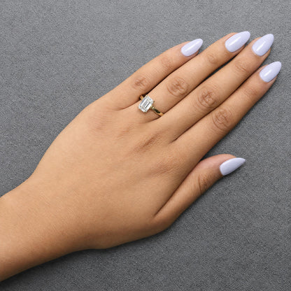 Close-up angled view of a yellow ring with a single emerald-shaped white gemstone and multiple small round white gemstones, shown worn on a model's finger.
