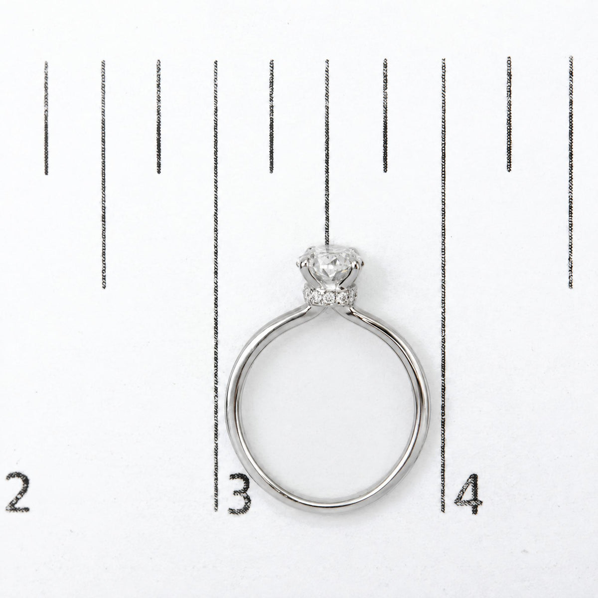 Top-down view of a white metal ring shown on a scale, featuring a central oval-shaped white gemstone in a 4-prong setting, surrounded by smaller round white gemstones in a prong setting around the band.