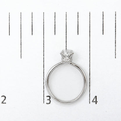 Top-down view of a white metal ring shown on a scale, featuring a central oval-shaped white gemstone in a 4-prong setting, surrounded by smaller round white gemstones in a prong setting around the band.