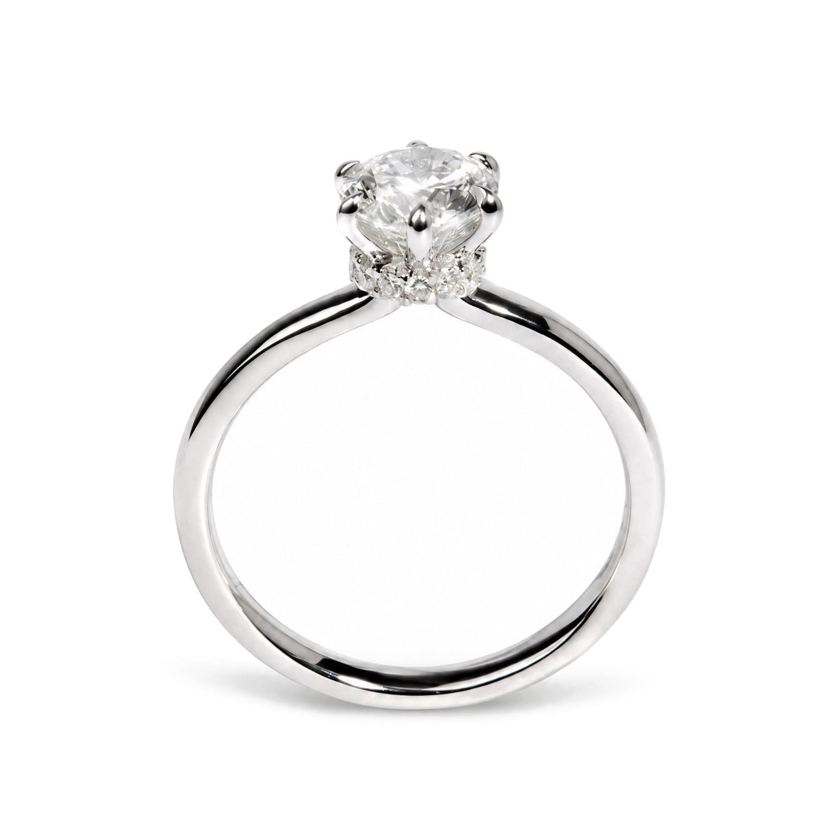 Close-up front view of a white metal ring featuring a single round white gemstone in a six-prong setting, surrounded by smaller round white gemstones in prong settings along the band.