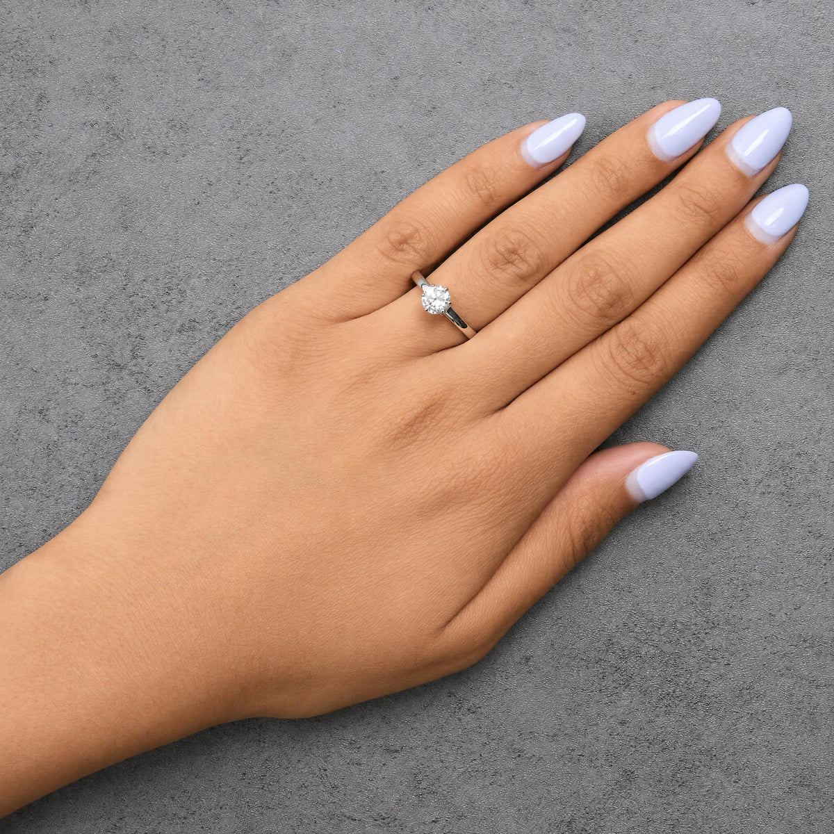 White metal ring with a round white gemstone in a 6-prong setting shown worn on a model's hand, top-down angle.
