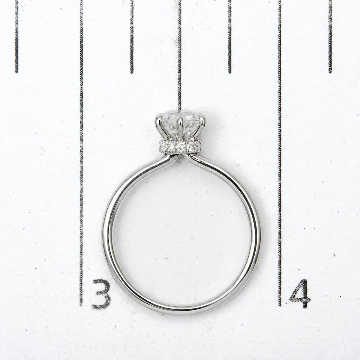 Top-down close-up view of a white metal ring with one round white gemstone in a six-prong setting and multiple smaller round white gemstones in prong settings, shown on a scale.