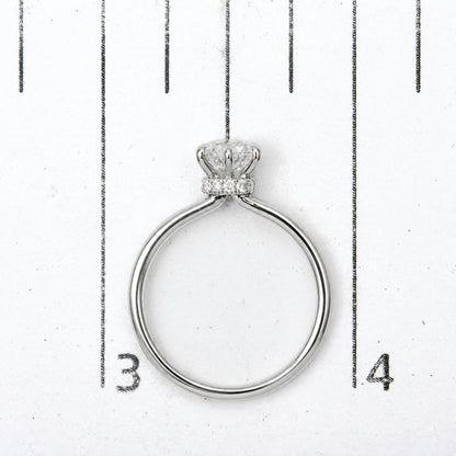 Top-down close-up view of a white metal ring with one round white gemstone in a six-prong setting and multiple smaller round white gemstones in prong settings, shown on a scale.