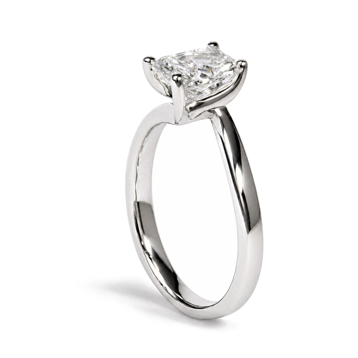 Close-up side angle of a white metal ring featuring one radiant-shaped white gemstone in a prong setting, shown on a white background.