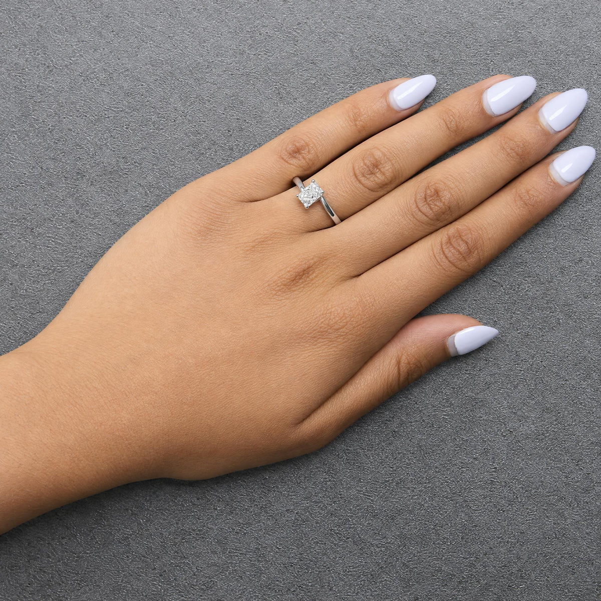 Close-up angled view of a white metal ring with a single radiant shape white gemstone in prong setting, shown worn on a model's finger with light purple nail polish.