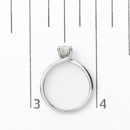 White metal ring with a single radiant-shaped white gemstone in a prong setting, shown in a close-up shot on a scale.