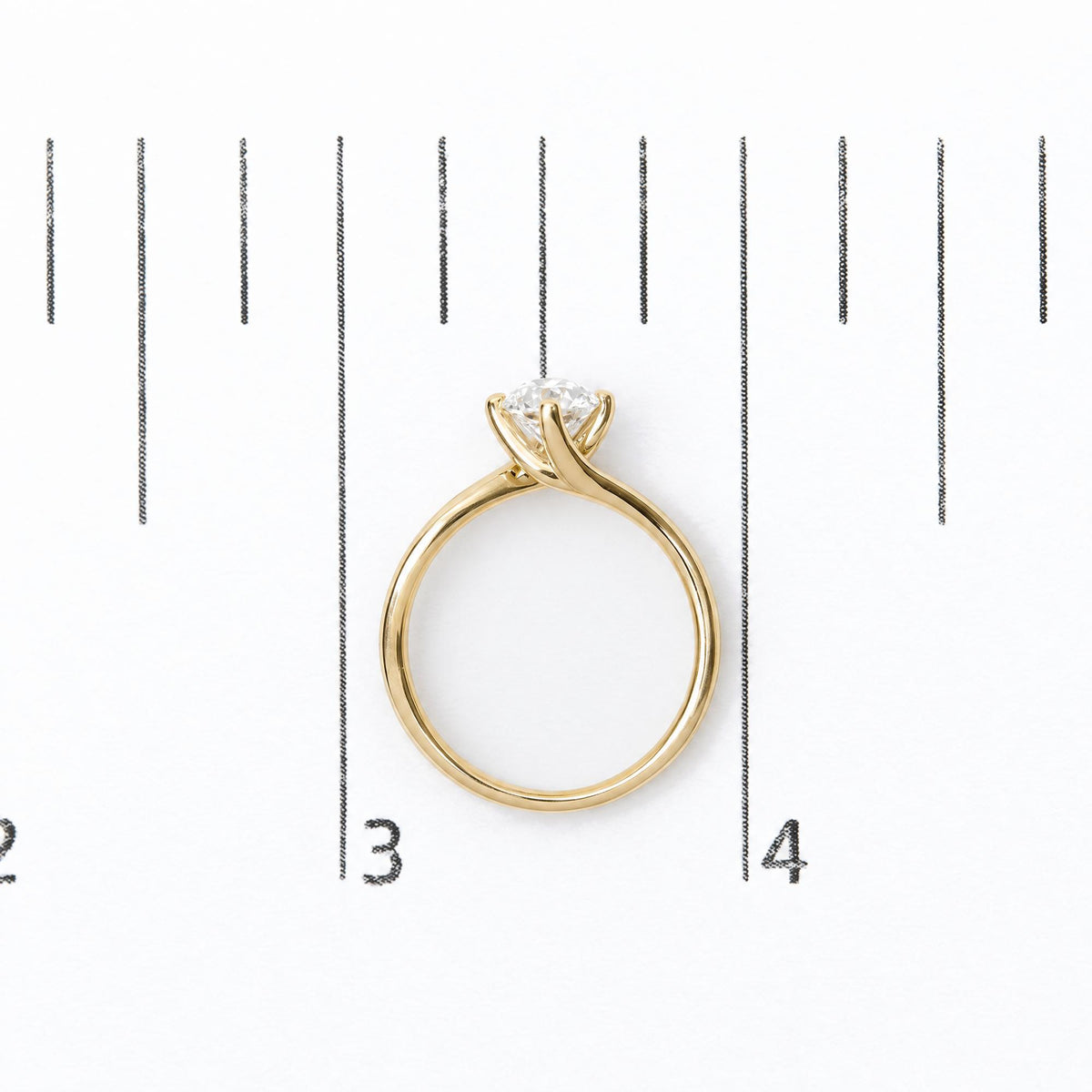 Close-up front view of a yellow metal ring with a single round white gemstone in a prong setting, shown on a measurement scale background.