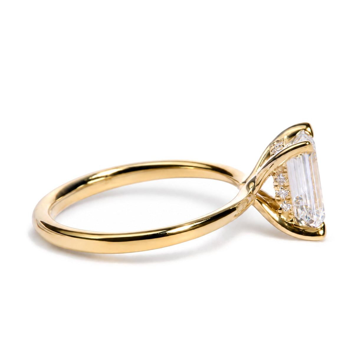 Close-up side angle of a yellow metal ring featuring one emerald-shaped white gemstone and surrounding round white gemstones in prong settings.